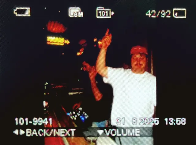 Camcorder shot of a DJ with one arm raised in a dark bar